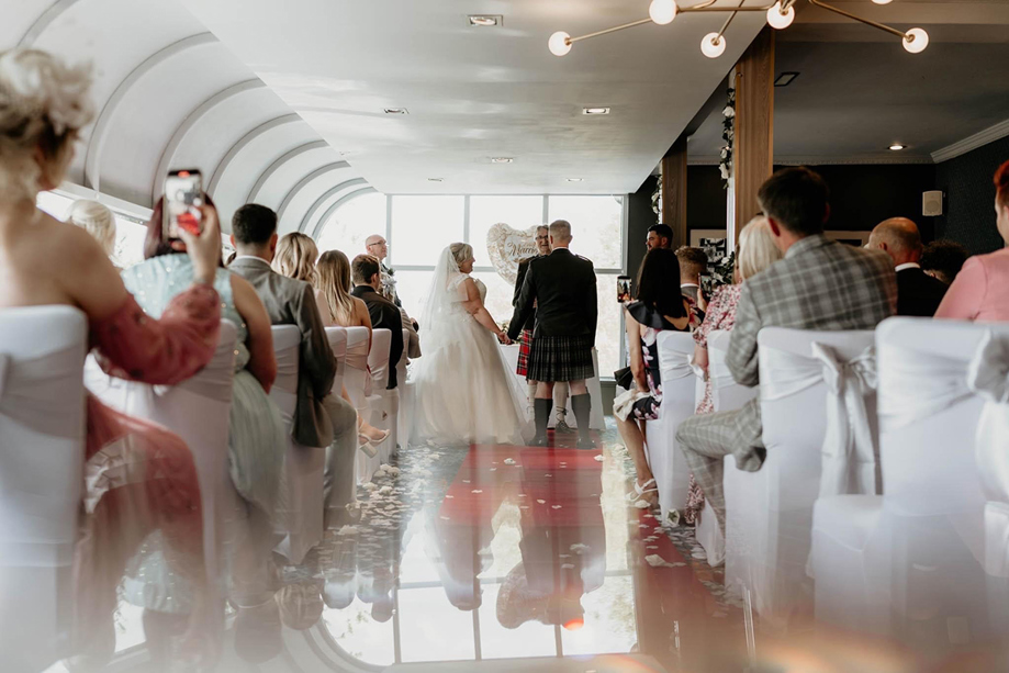 Bride and groom stand at altar holding hands during ceremony, while guests look on from white chairs.